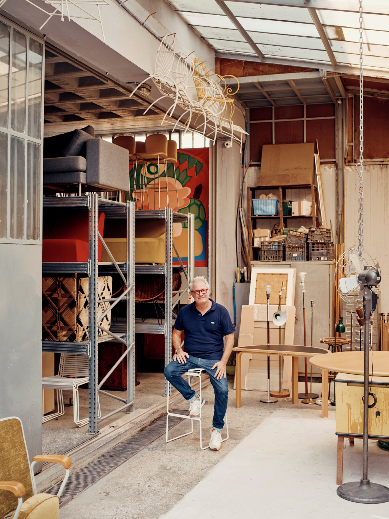 Alketas Pazis sitting on white chair in warehouse showroom filled with vintage furniture and industrial shelving