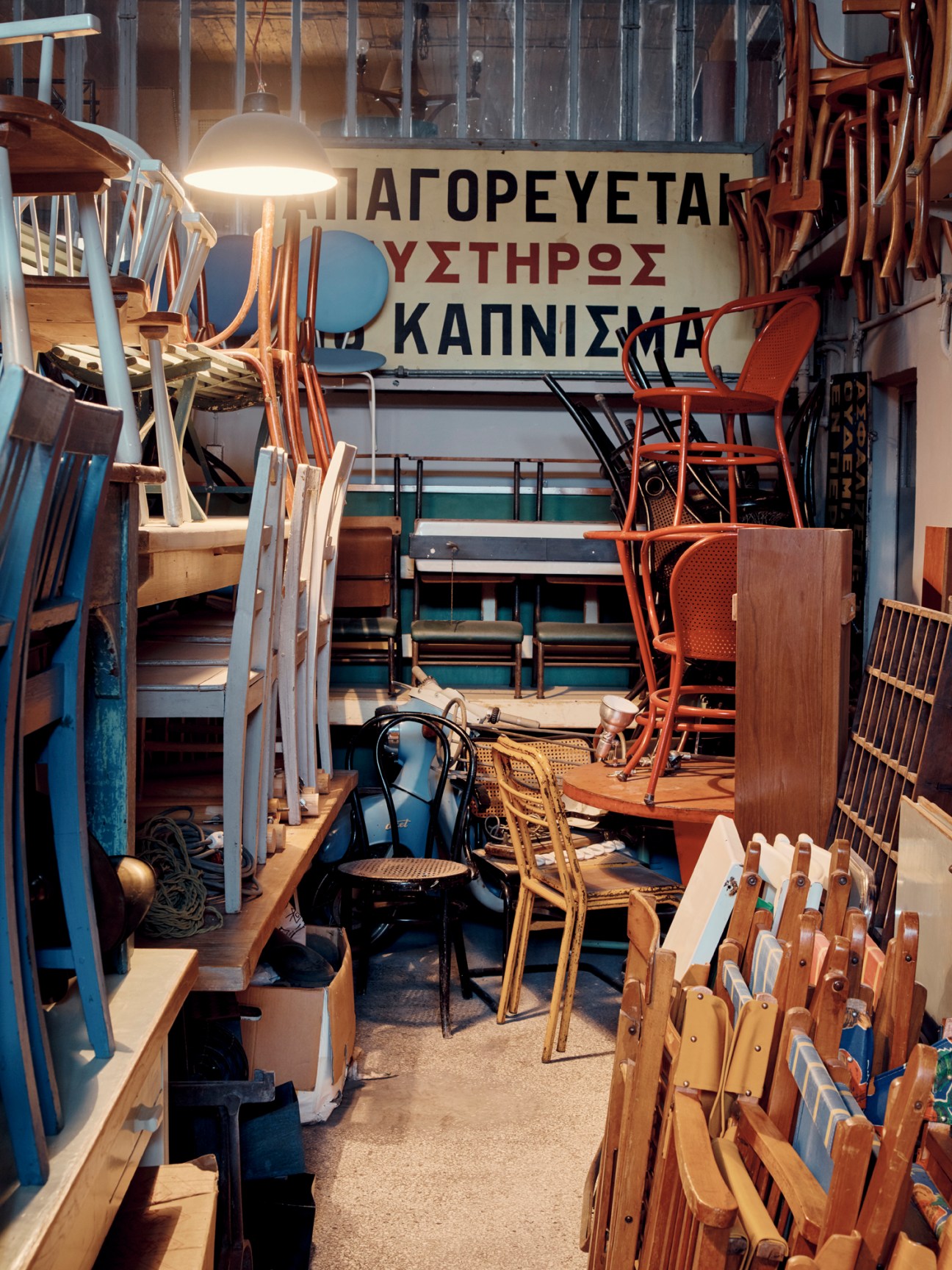 Warehouse space filled with stacked vintage chairs and furniture with Greek signage on the wall