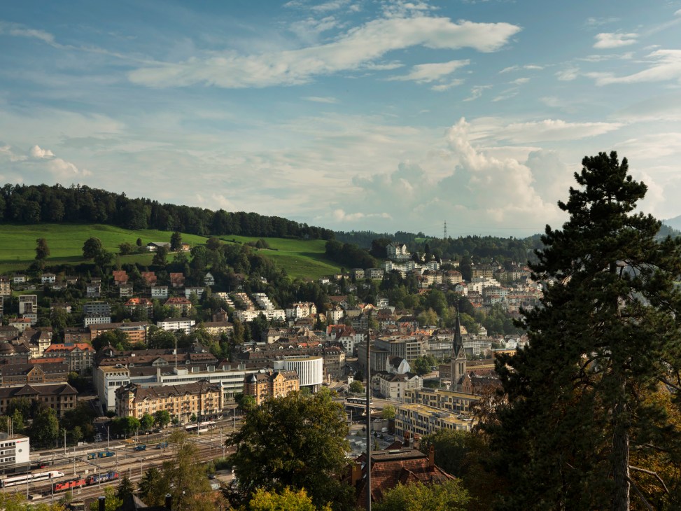Panoramic view of St Gallen with rolling green hills, residential buildings, and forested mountains in background