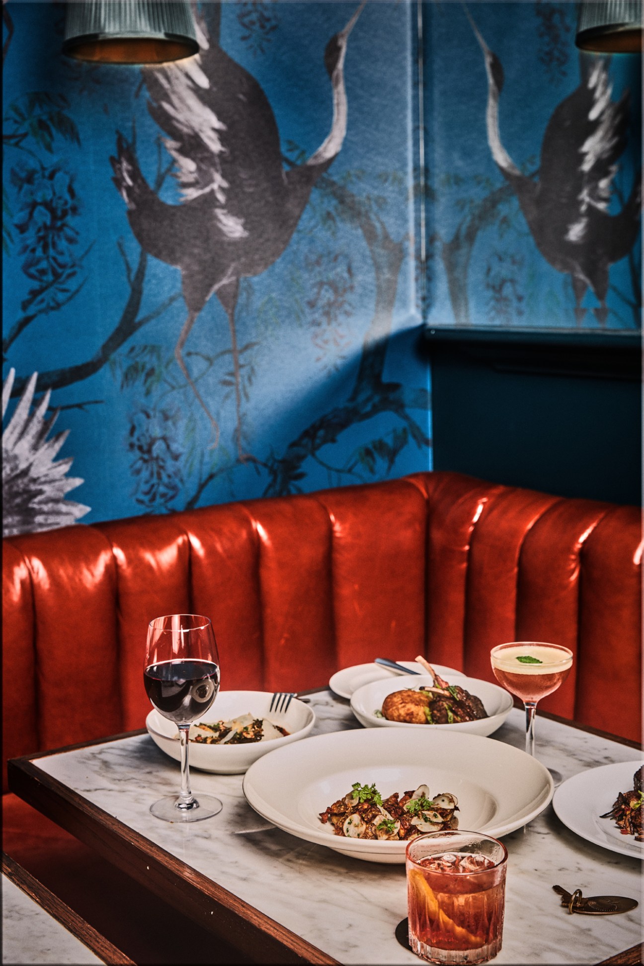 Restaurant table with plated dishes and cocktails against red banquette seating and blue crane wallpaper