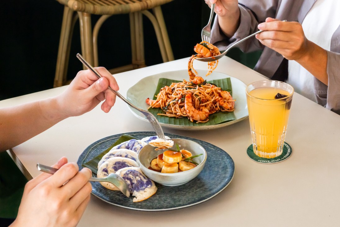 Two people eating Asian dishes with chopsticks and forks at a table with noodles, dumplings and orange juice