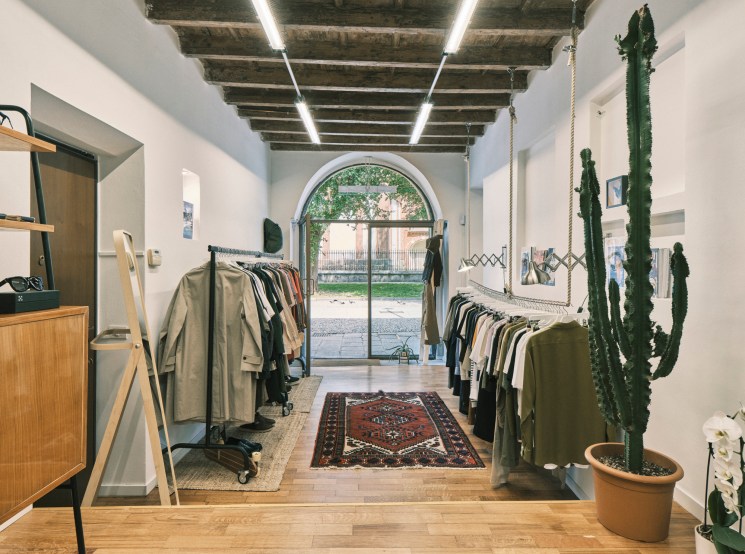 Dictionary clothing shop interior in Milan with minimal white walls, wooden flooring, clothing racks, potted cacti, and arched doorway opening to courtyard.