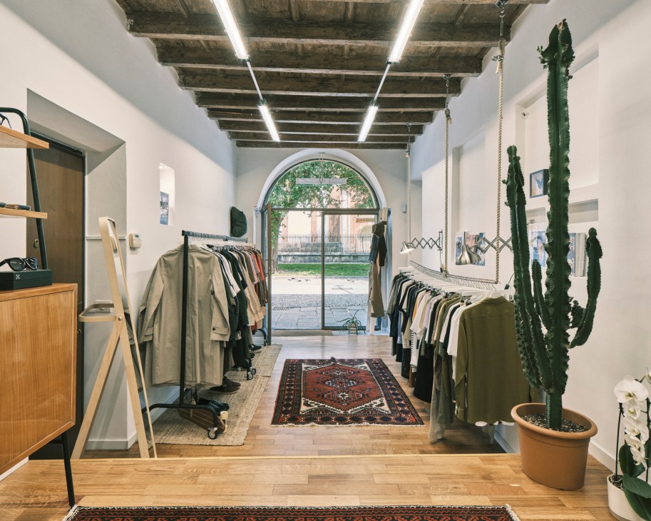 Dictionary clothing shop interior in Milan with minimal white walls, wooden flooring, clothing racks, potted cacti, and arched doorway opening to courtyard.