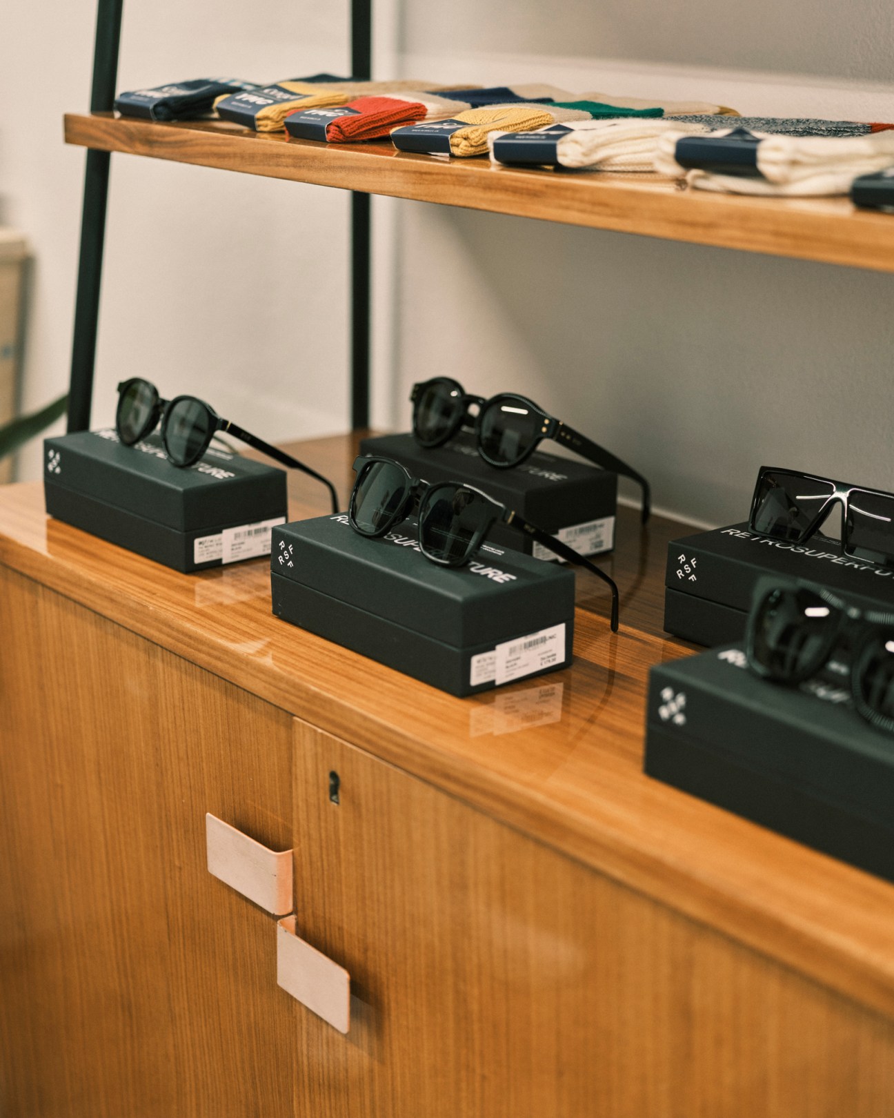 Sunglasses by Retrosuperfuture displayed on black boxes on wooden shelving at Dictionary Milan, with folded items visible on shelf above.