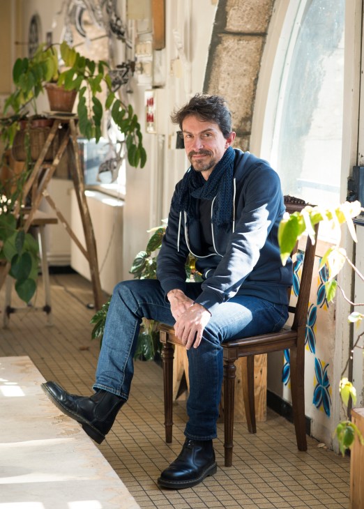 Man seated on wooden chair in plant-filled passageway with natural light from window