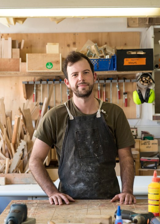 Man wearing work apron standing at wooden workbench in organized workshop with tools and materials.