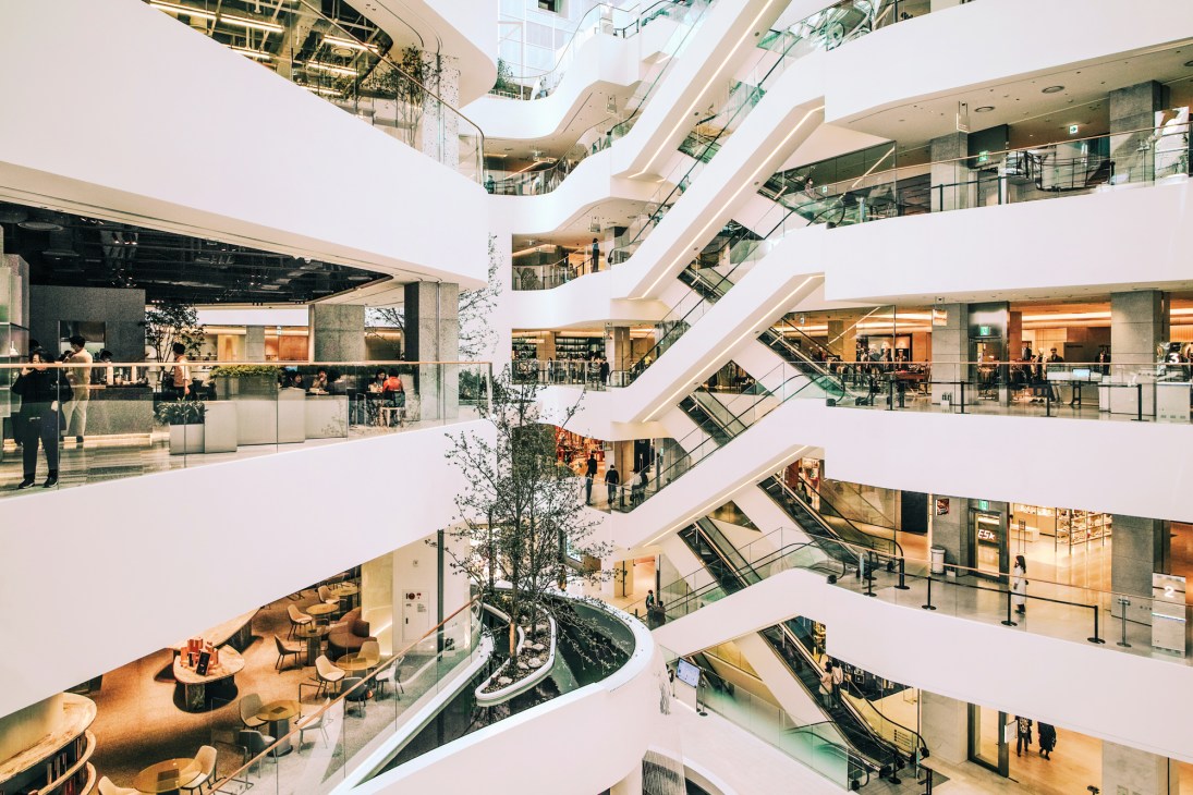 Multi-level shopping mall interior with white curved balconies, escalators, and natural skylighting.