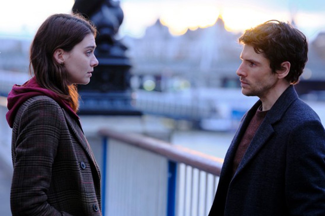 Two people face each other on a bridge with a blurred cityscape at dusk in the background.
