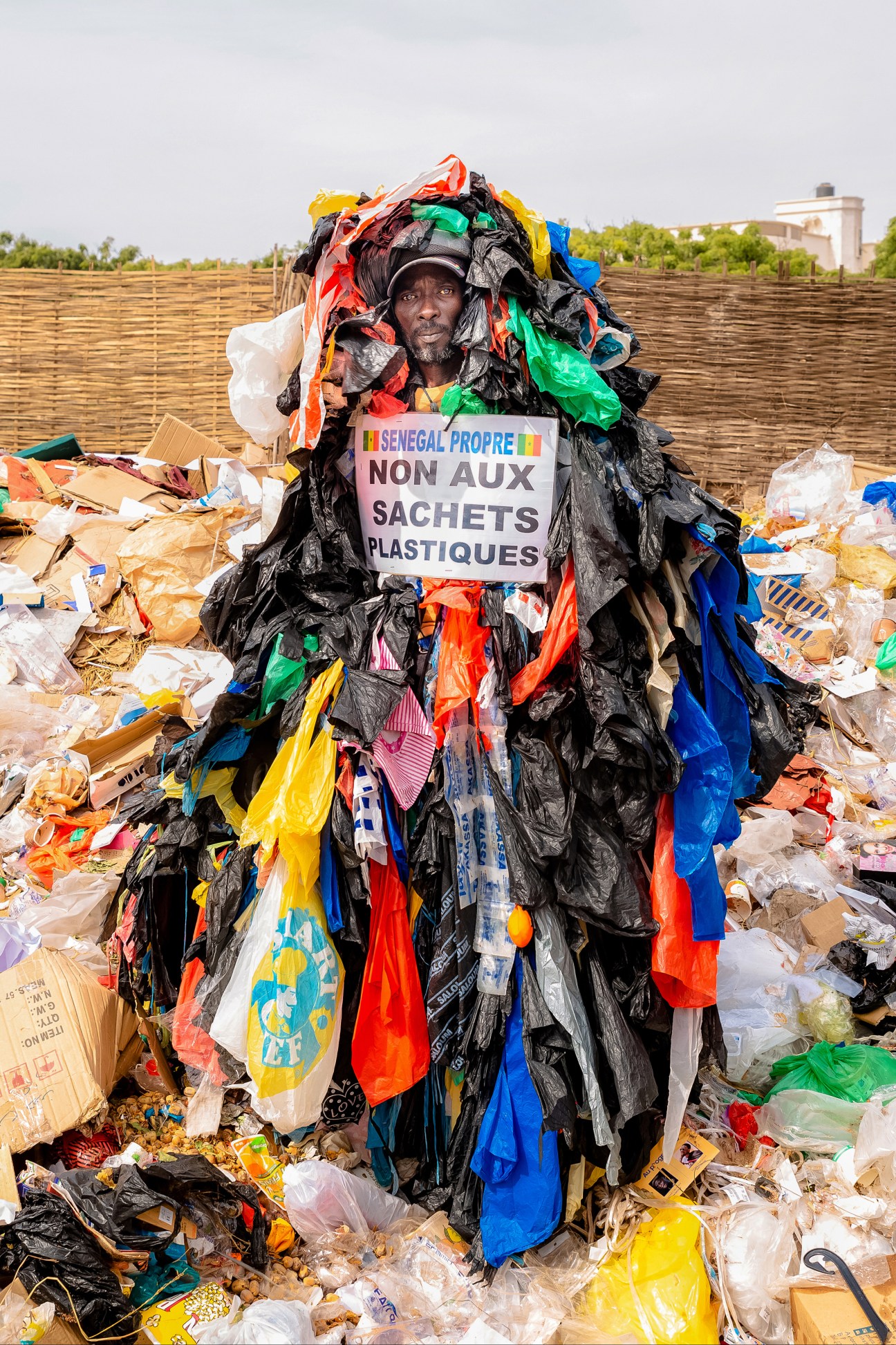 Modou Fall covered in colorful plastic bags holds sign reading 'Senegal Propre Non Aux Sachets Plastiques' amid waste