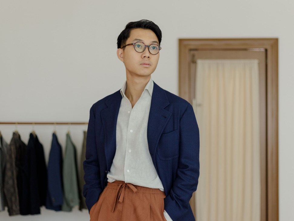 Asian man in navy blazer and white shirt stands in tailoring studio with suits hanging in background
