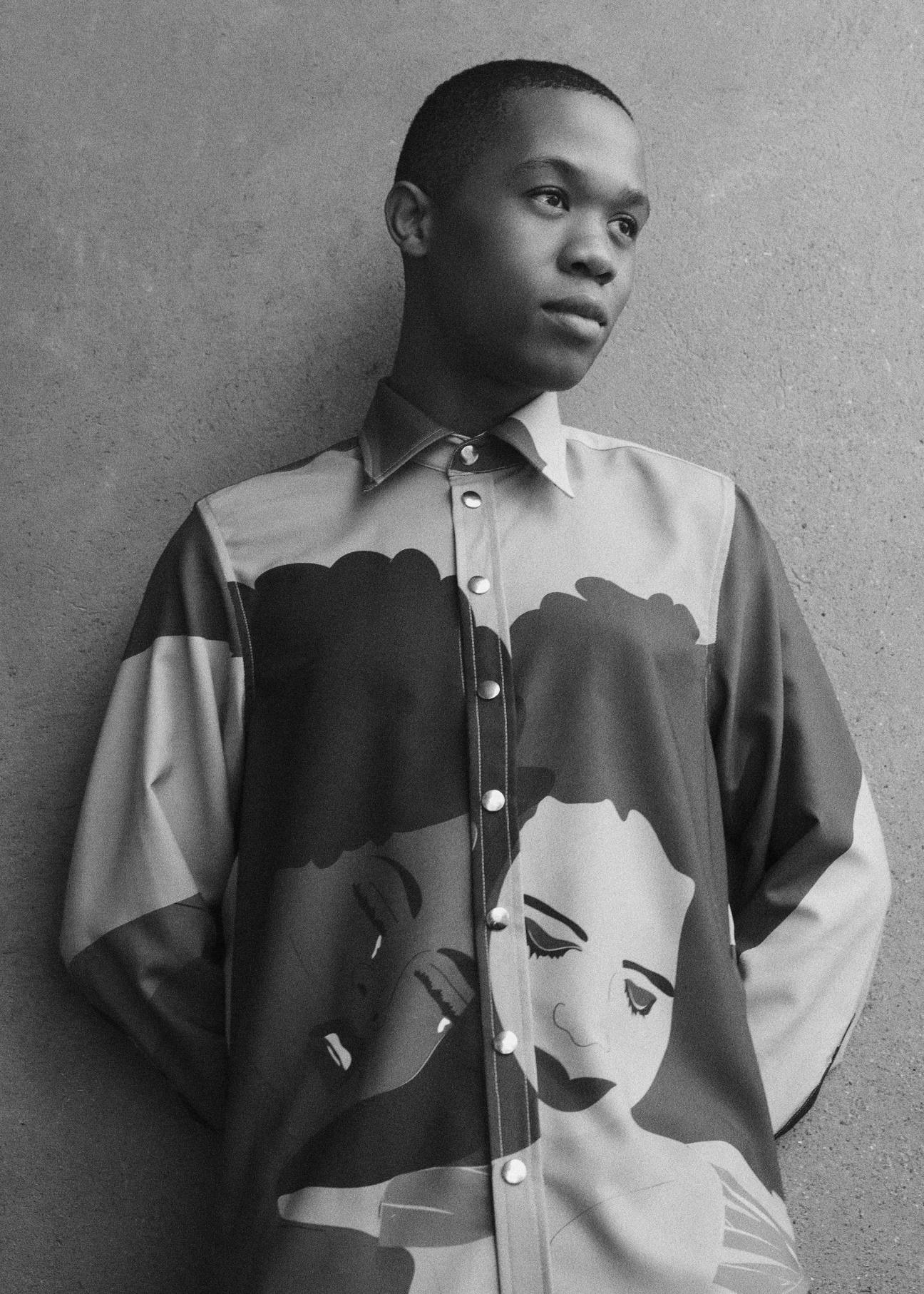 Thebe Magugu, a Black man in a patterned button-up shirt with illustrated faces, standing against a plain wall
