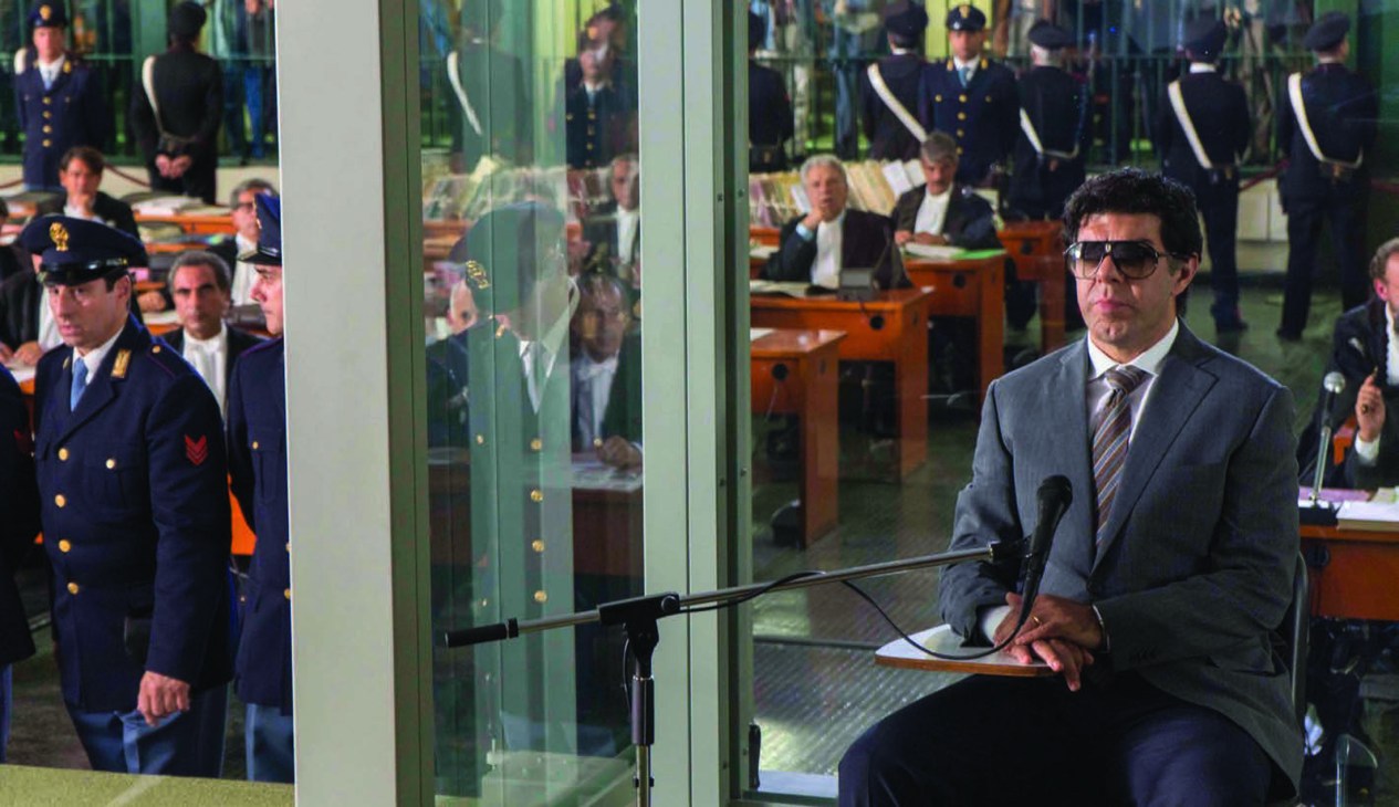 A man in a gray suit and sunglasses sits at a microphone in a courtroom with military officers in the background.