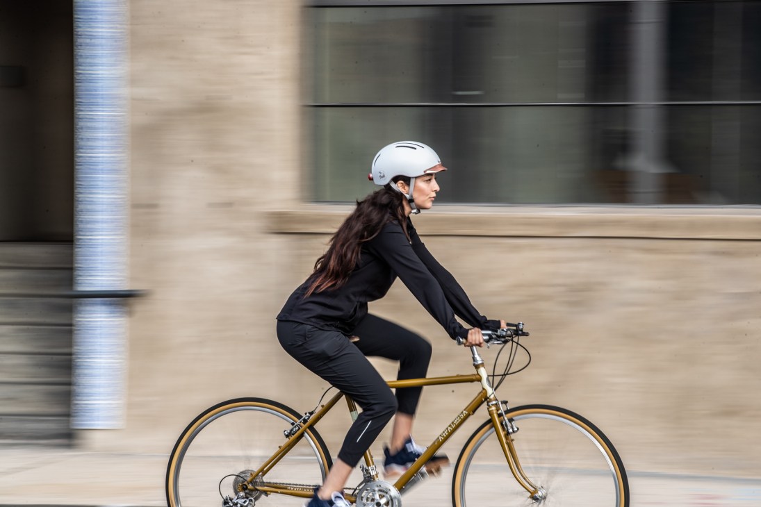 Woman cycling on urban street wearing white Thousand helmet and black athletic clothing.
