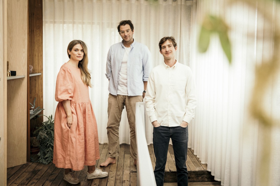 Three people standing together in a modern interior space with wooden floors and white curtains