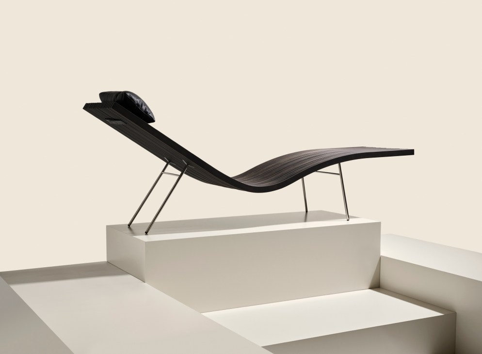 Black modern chaise lounge with curved seat and headrest pillow on thin metal legs, displayed on a white platform pedestal against a neutral background.