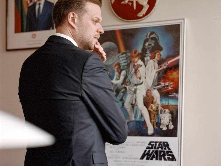 Man in dark suit standing in office with Star Wars poster and coat of arms on wall