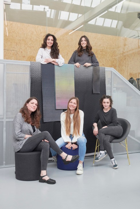 Five female staff members pose together in a modern office space with cork walls and skylight windows.
