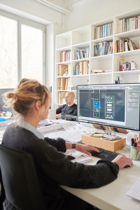 Two designers work at desks in a bright office space with white shelving and a computer displaying architectural drawings.