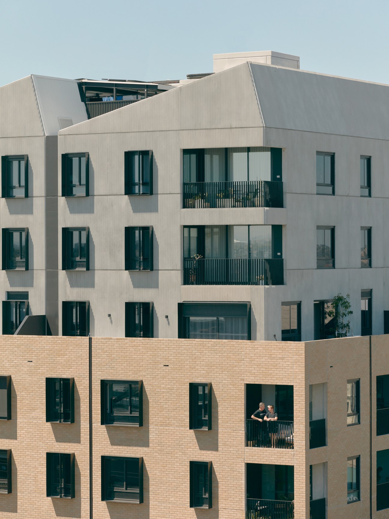 Modern residential apartment building with cream corrugated metal upper levels and warm brick lower facade featuring dark-framed windows.