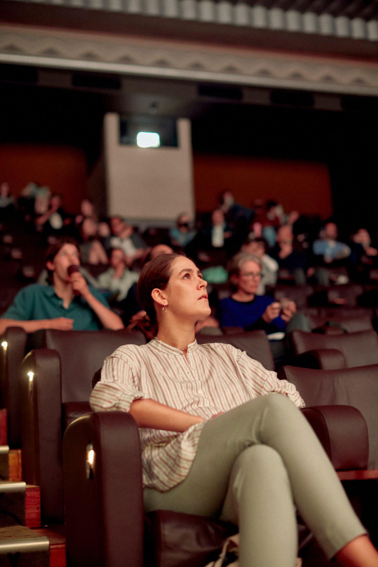 Woman in striped shirt looking up thoughtfully while seated in cinema auditorium with other audience members