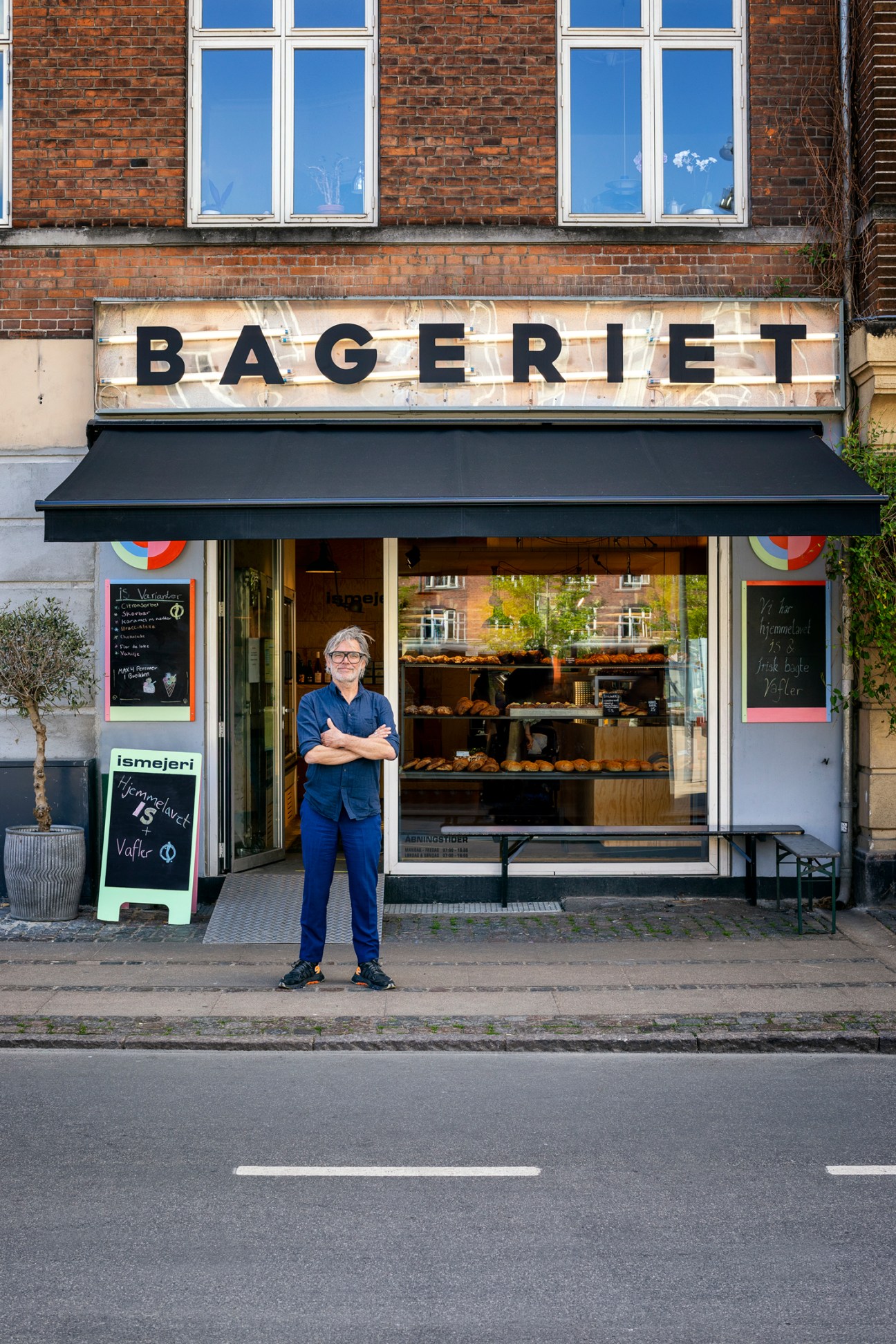 Man in blue shirt and glasses standing with arms crossed outside brick building bakery storefront