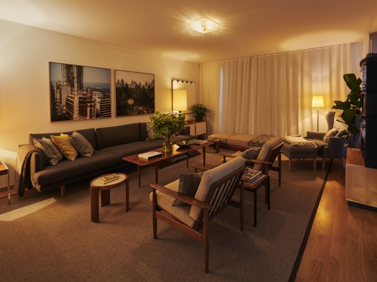 A warmly lit modern living room with a dark sofa, mid-century wooden chairs, indoor plants, and city photography on the wall.