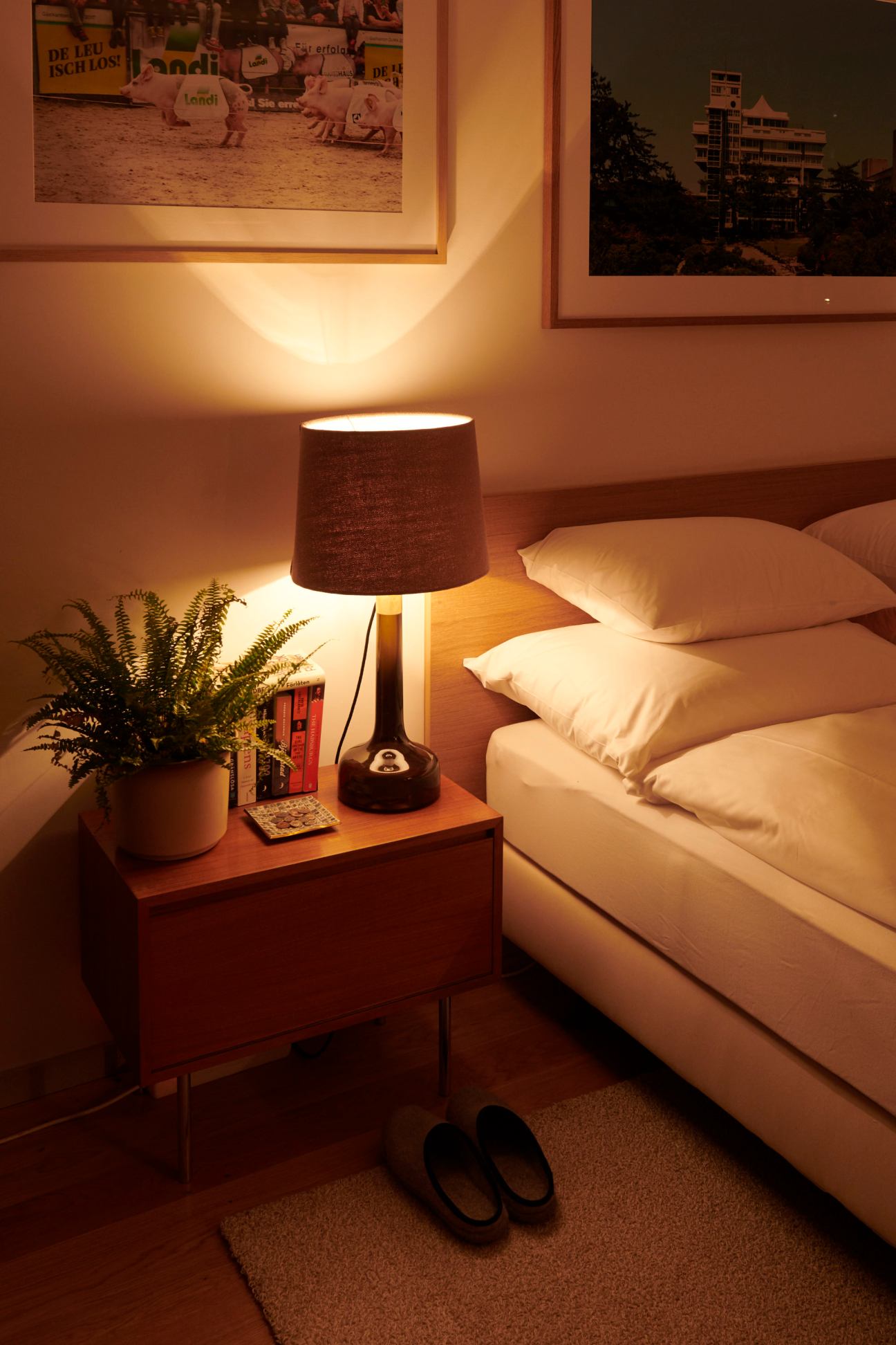 Warm bedside scene with table lamp casting golden light, white pillows, wooden nightstand, plant and slippers on floor