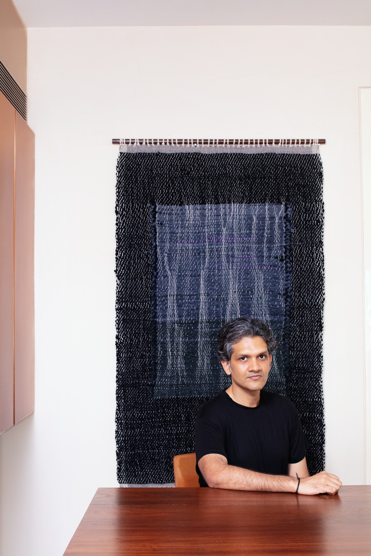 Collector Udit Bhambri poses in front of a woven tapestry artwork by Monika Correa featuring black and blue textile patterns.