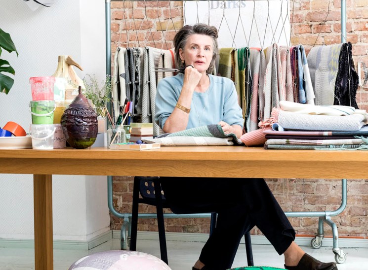 Hella Jongerius sitting at her desk in her Berlin studio workspace with clothing rack and exposed brick wall behind