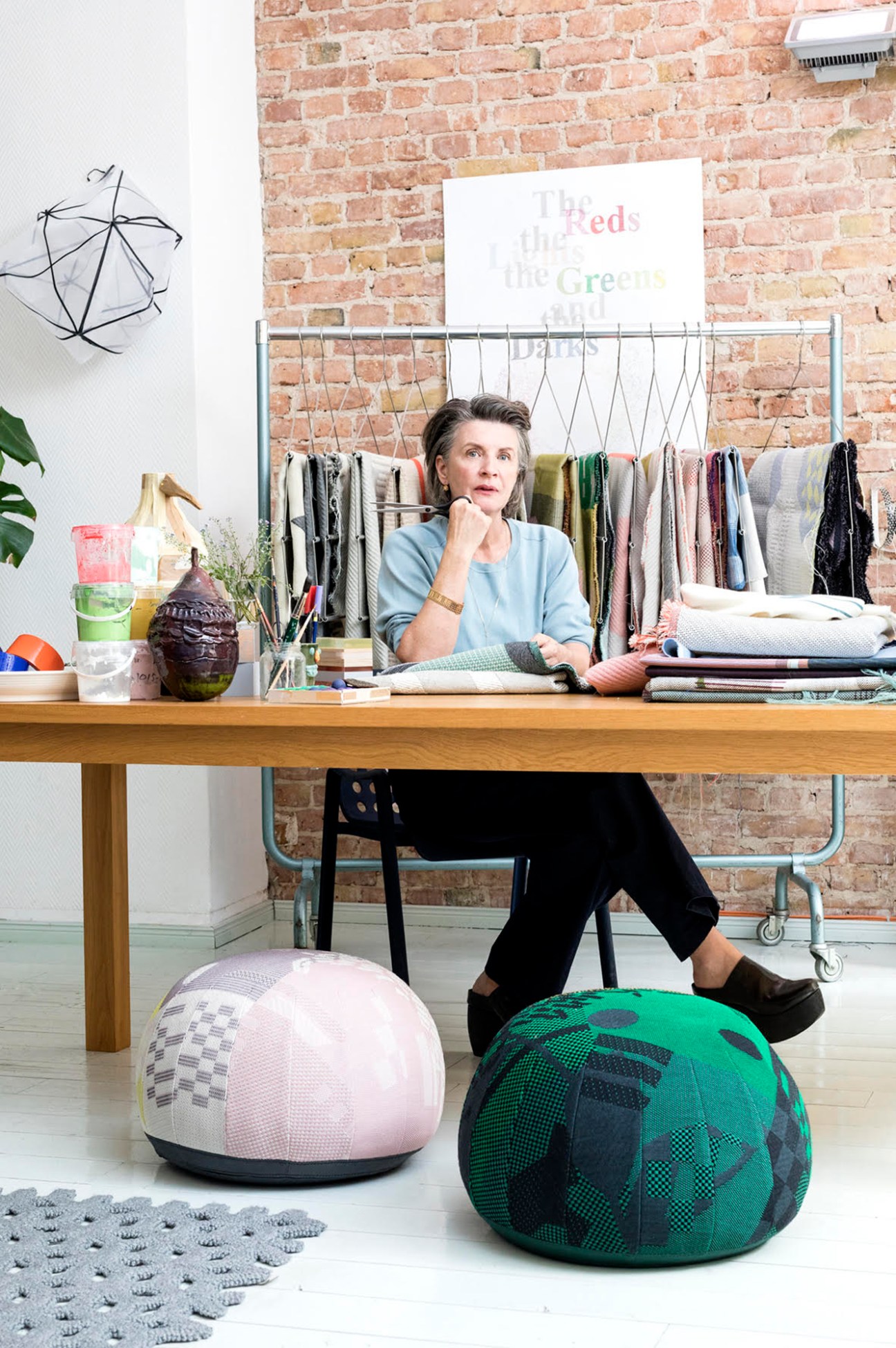 Hella Jongerius sitting at her desk in her Berlin studio workspace with clothing rack and exposed brick wall behind