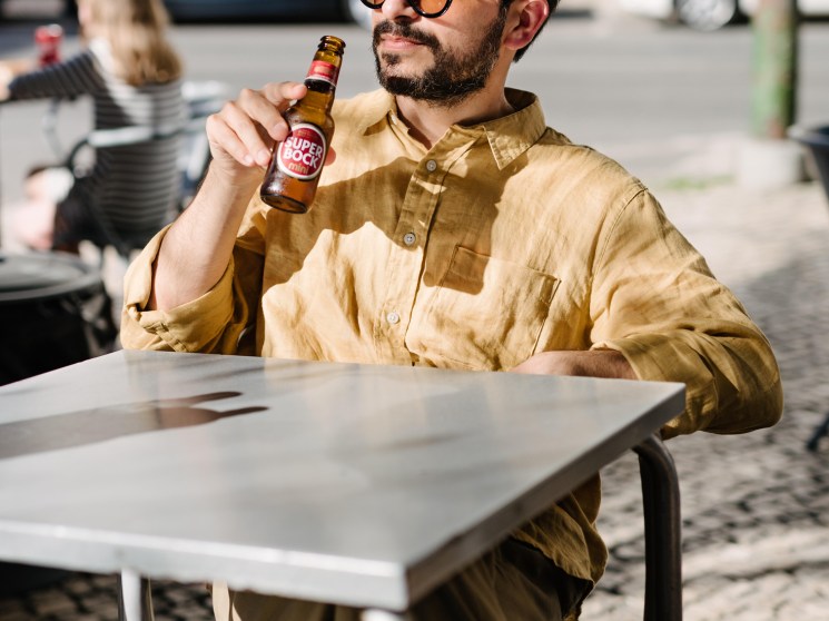 Man in sunglasses and yellow shirt holding a small brown Super Bock beer bottle at an outdoor table