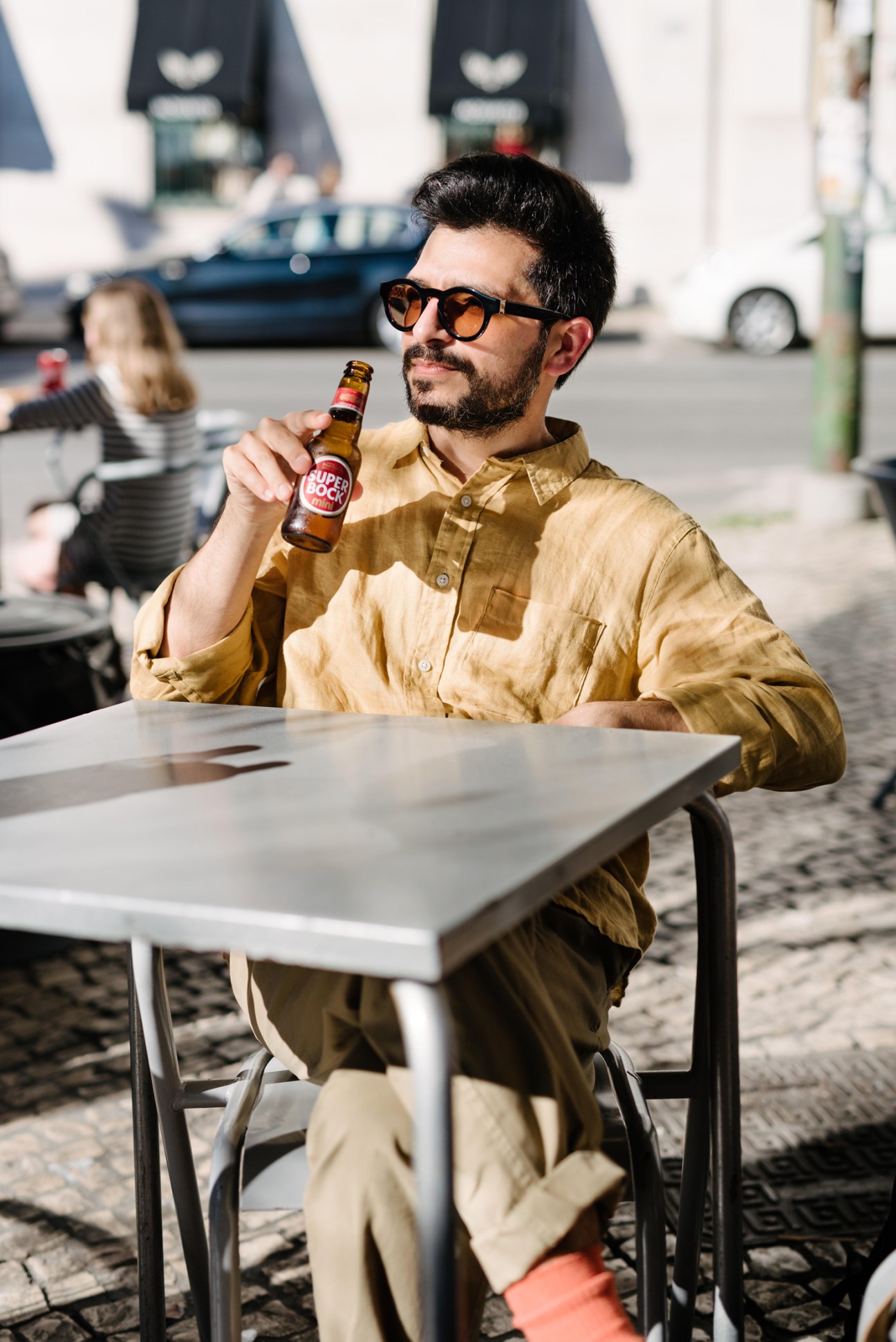 Man in sunglasses and yellow shirt holding a small brown Super Bock beer bottle at an outdoor table