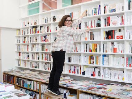 Woman reaching for books on tall white shelves in École de la Librairie bookshop classroom