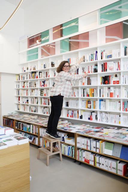 Woman reaching for books on tall white shelves in École de la Librairie bookshop classroom