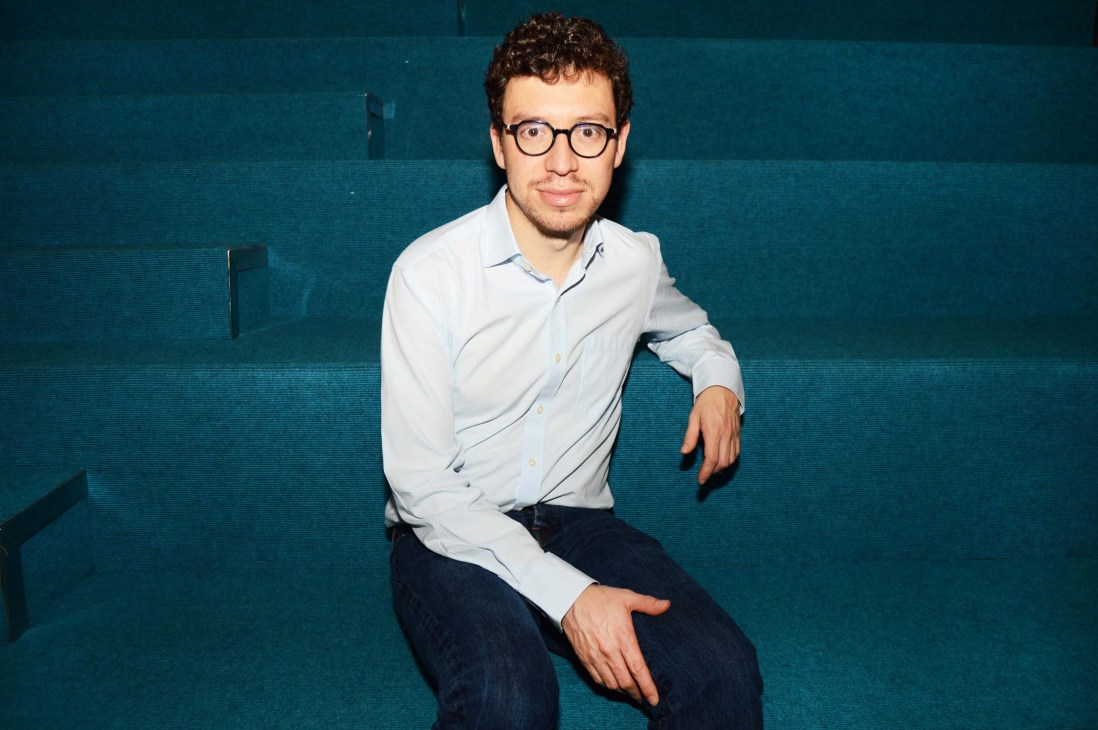 Man wearing light blue shirt and glasses poses against teal blue background.