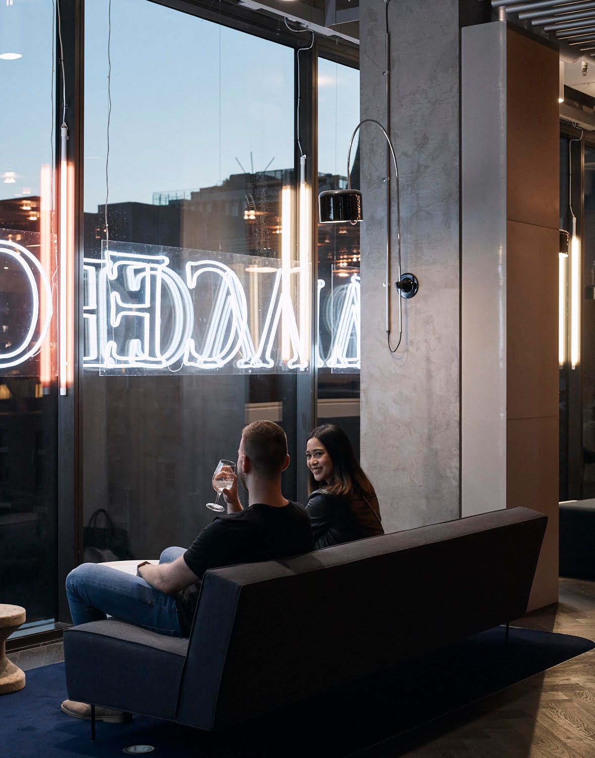 A couple sitting on a modern couch sharing drinks in an upscale cinema lounge with neon signage