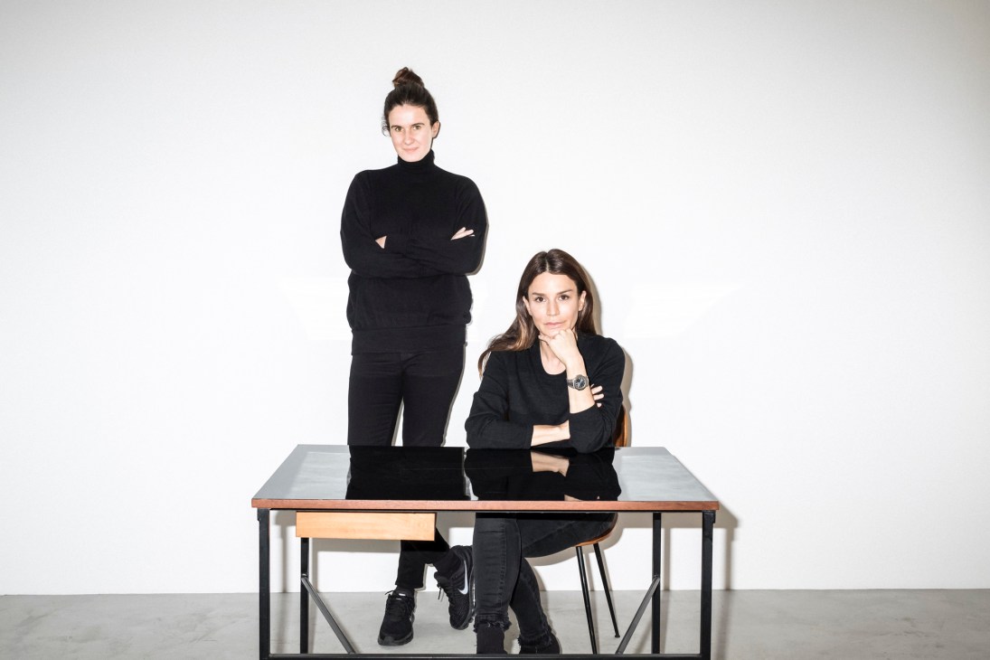 Two women in black clothing posed at a modern table with metal frame against white background