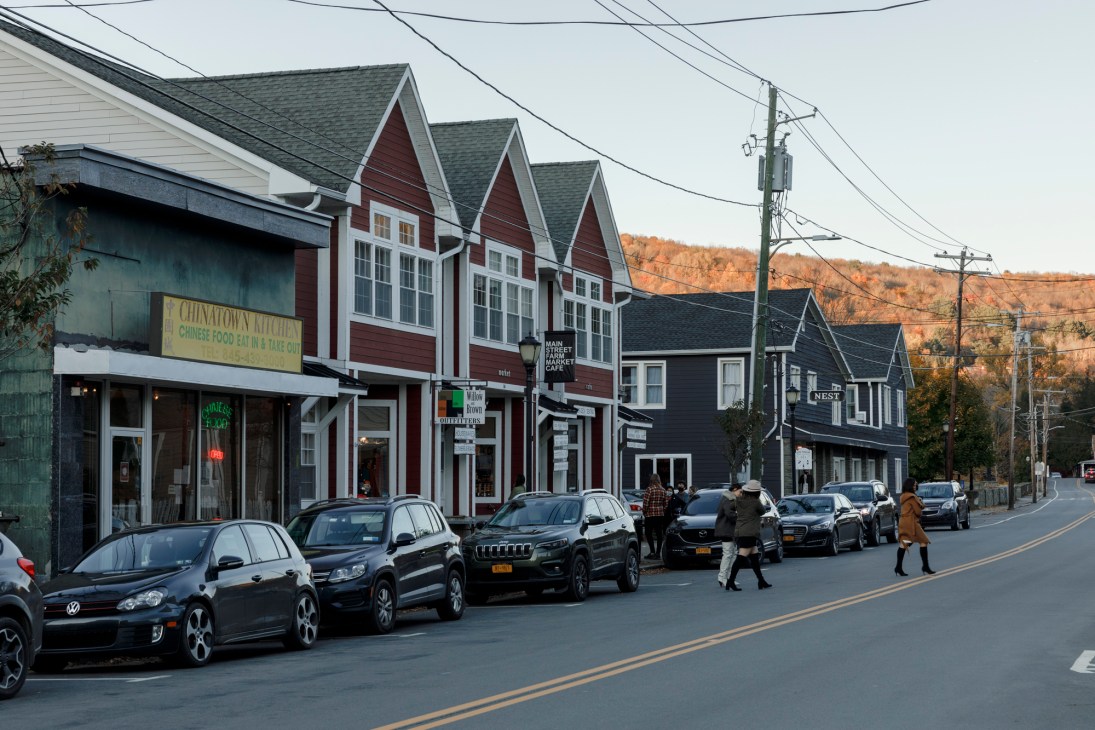 Row of traditional residential buildings with red and dark siding, storefronts at street level, parked cars, and autumn foliage in background hills.