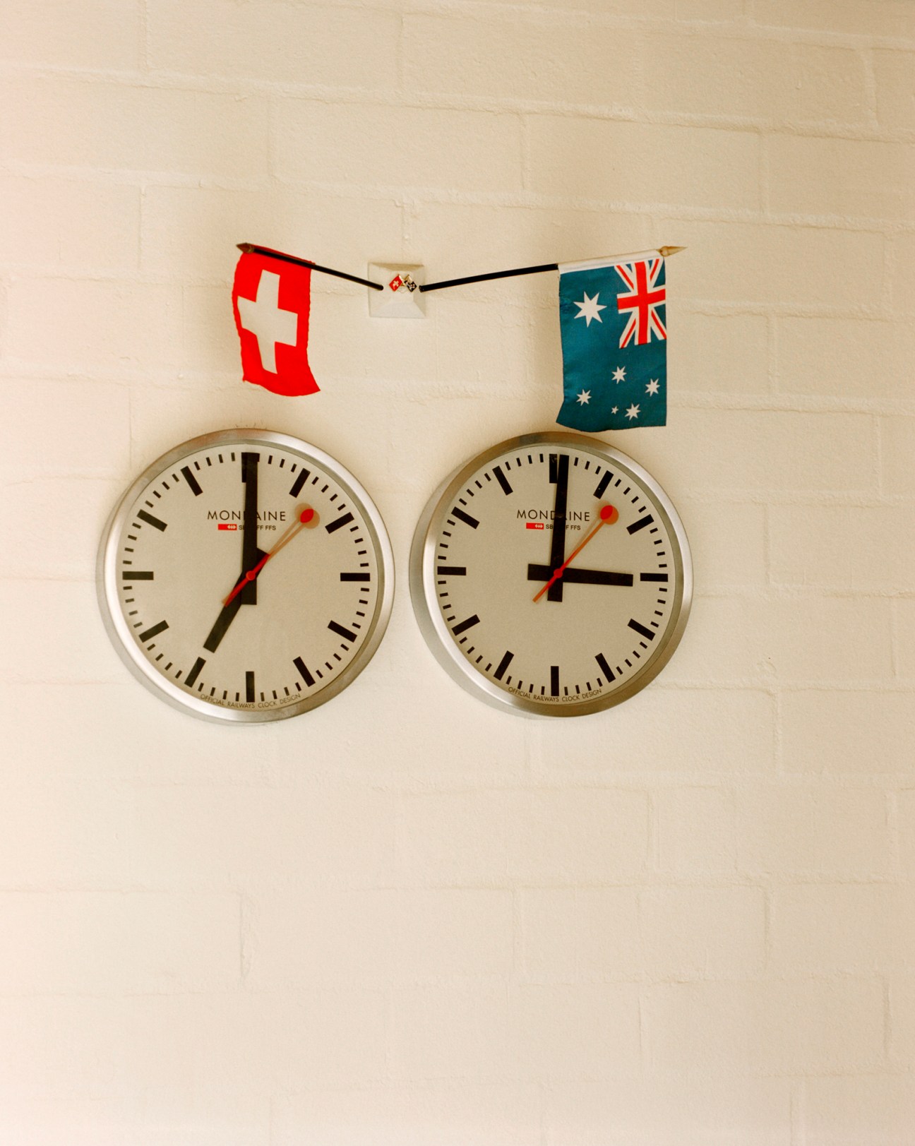 Two Mondaine clocks showing different times mounted on white wall with Swiss and Australian flag bunting above