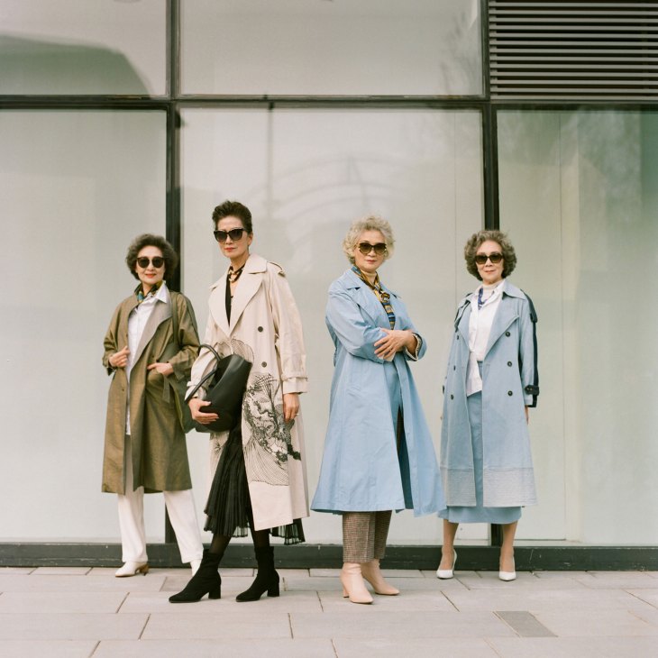 Four elderly women in trench coats and sunglasses pose against a modern glass building in Beijing