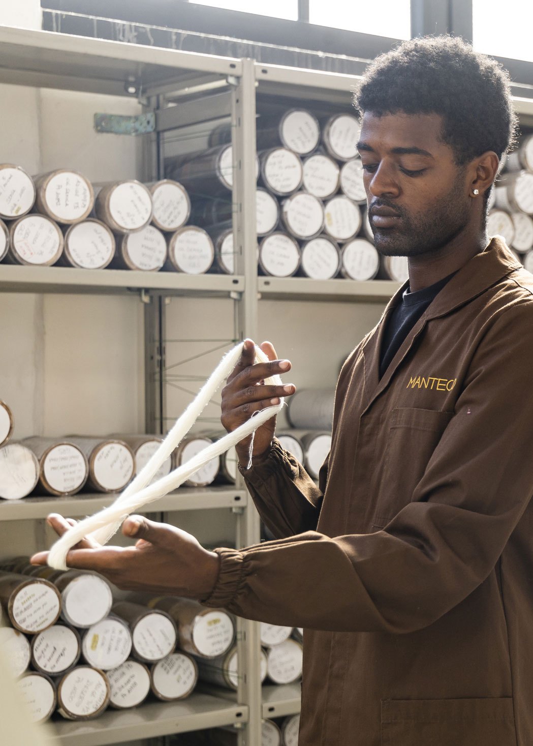 Worker at Manteco factory examining white yarn with shelves of fabric rolls in background