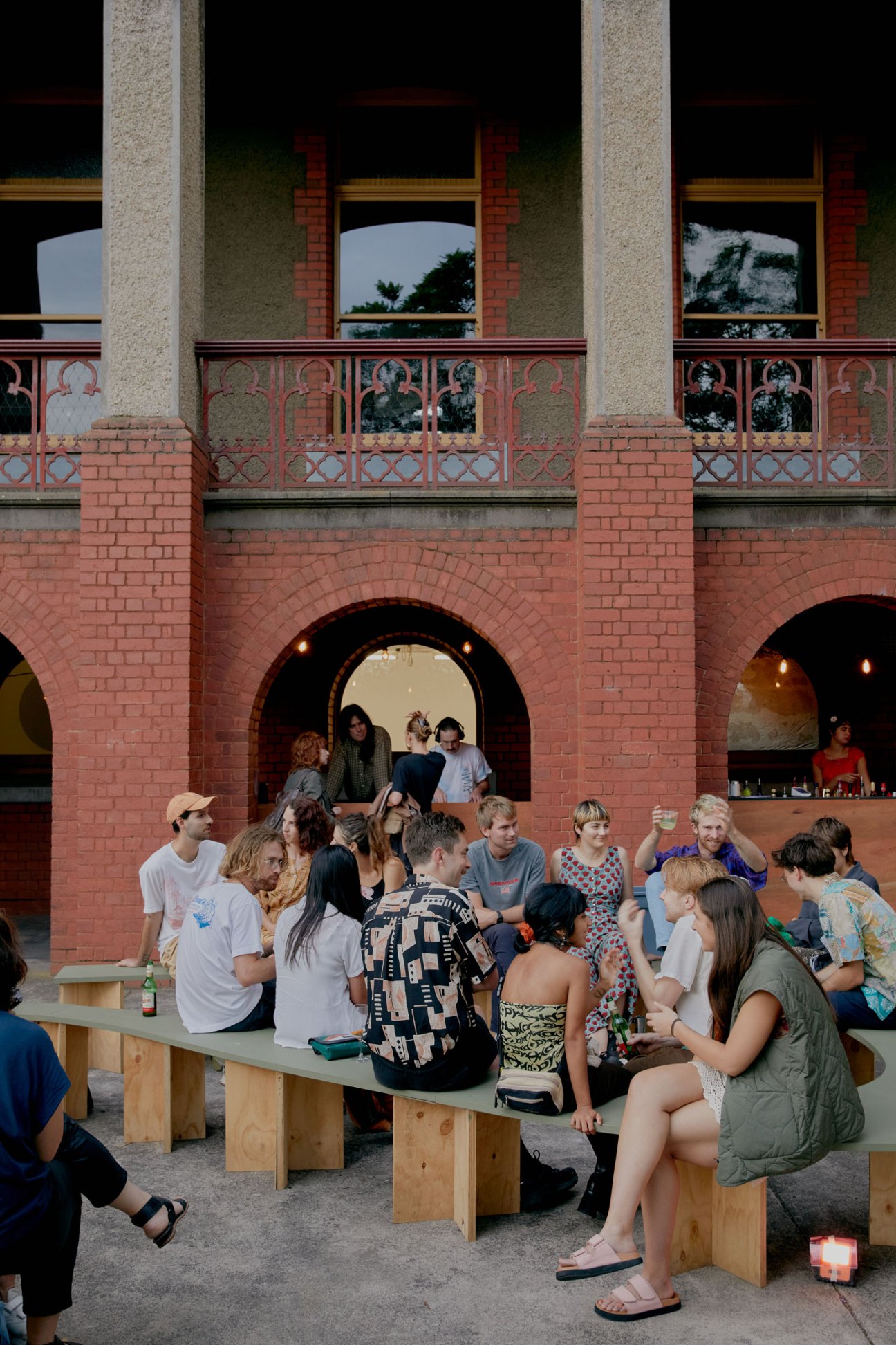 People sitting at wooden benches outside historic brick building with arched openings and ornate balcony