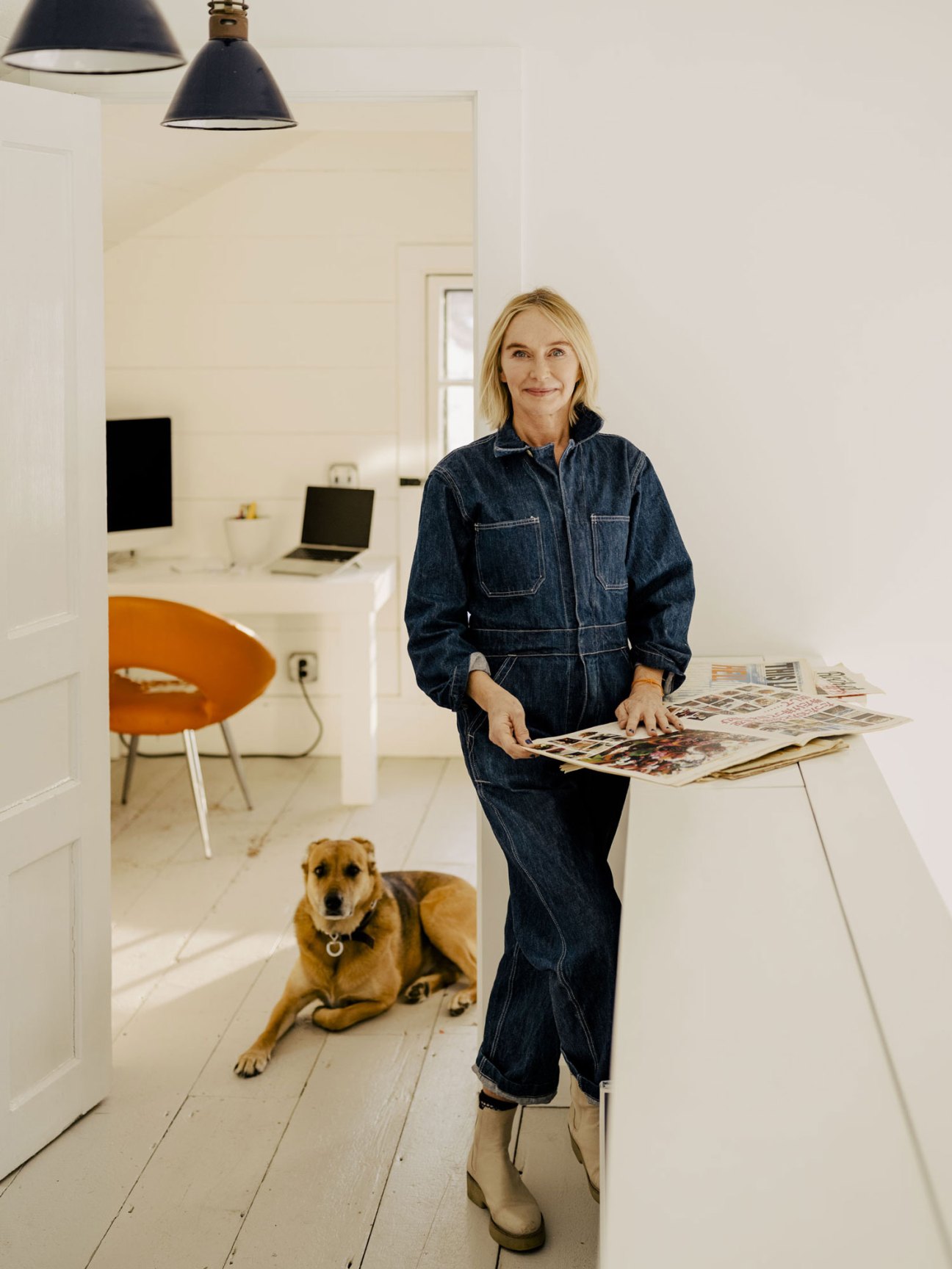 Woman in denim jumpsuit standing at white kitchen island with magazines and dog lying on floor