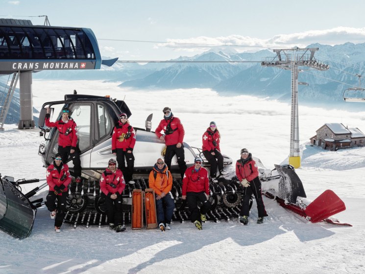 Nine avalanche security team members in red winter gear posing with equipment around a snowcat at Crans Montana ski resort