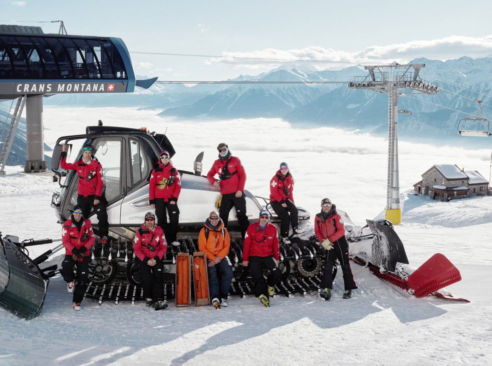 Nine avalanche security team members in red winter gear posing with equipment around a snowcat at Crans Montana ski resort