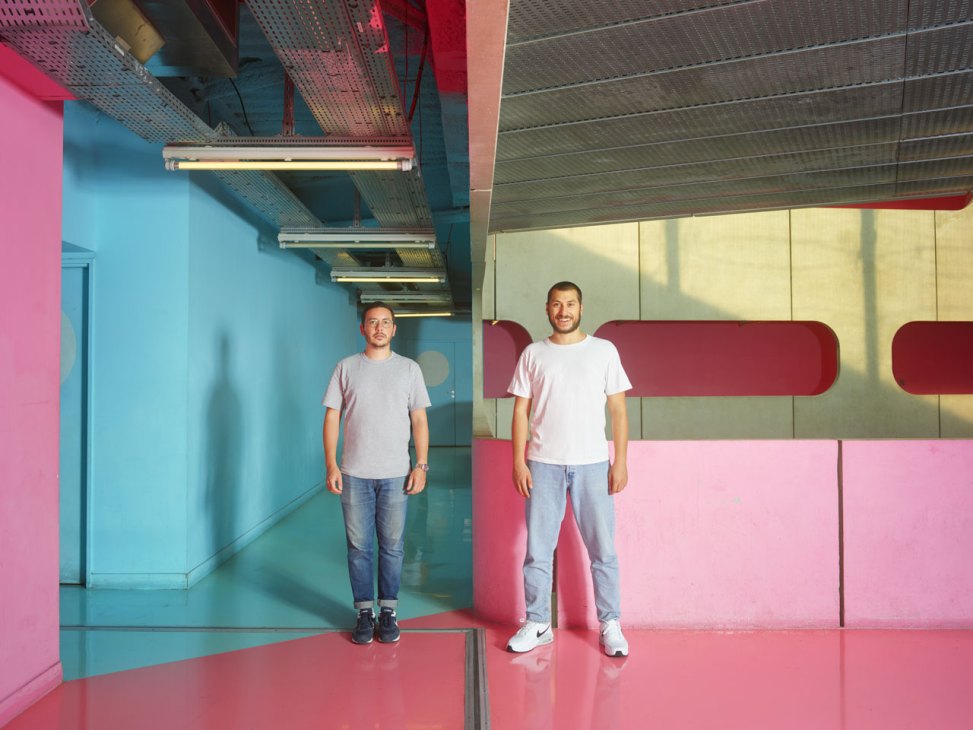 Two men standing in a modern corridor with colorful pink, blue and yellow walls and fluorescent lighting.