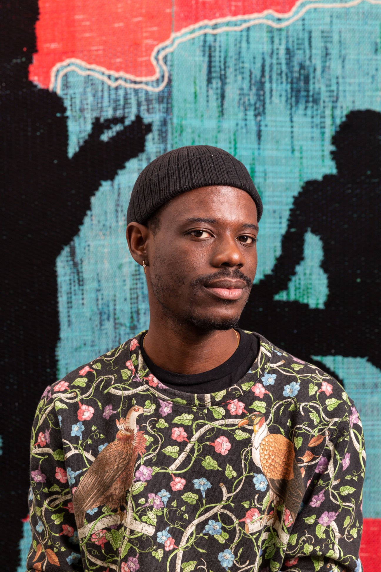Portrait of a Black man wearing a knit beanie and floral patterned shirt against a colorful mural background