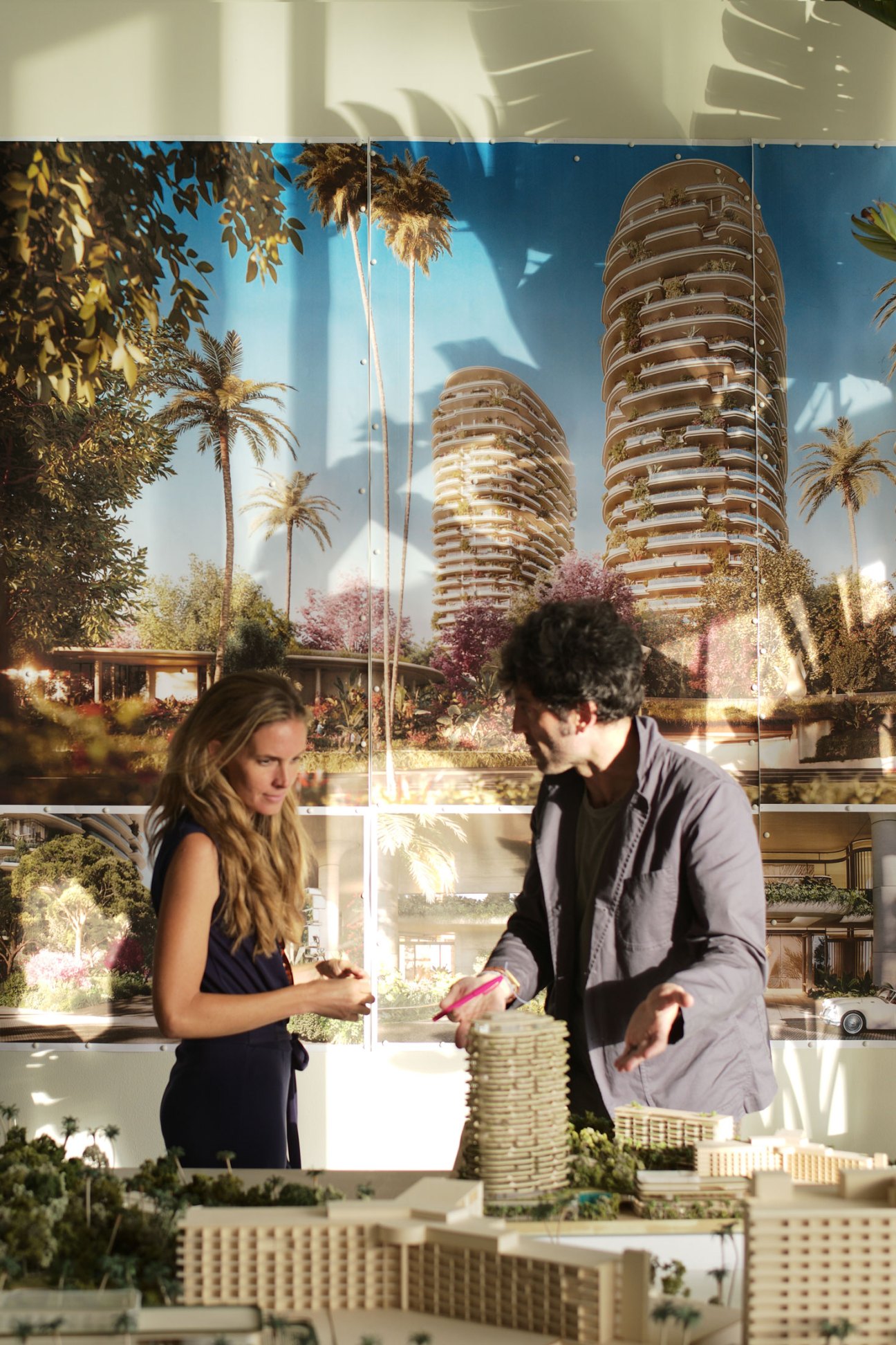 Two people examining an architectural model of modern high-rise towers with tropical landscaping in a presentation room
