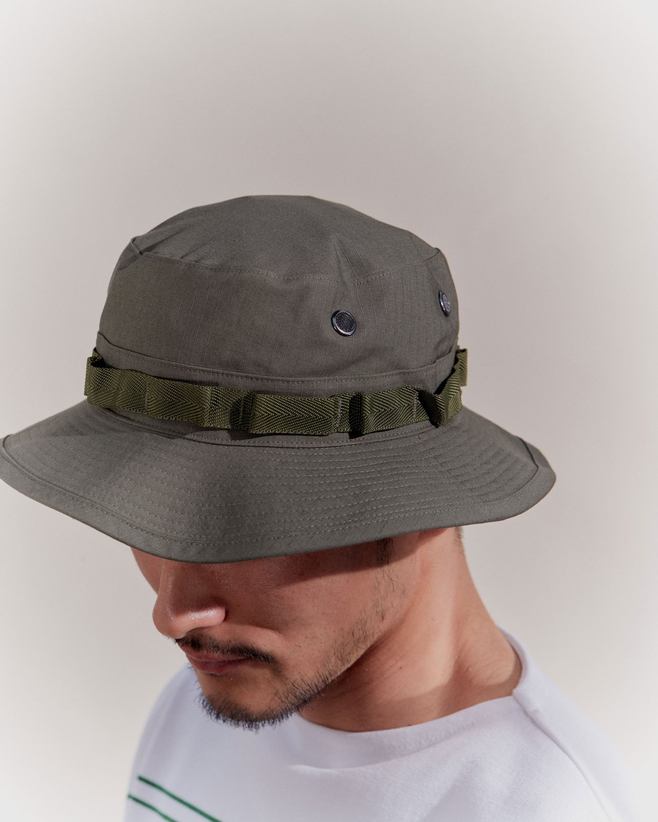 Man wearing olive green bucket hat with woven band detail and white striped shirt against neutral background