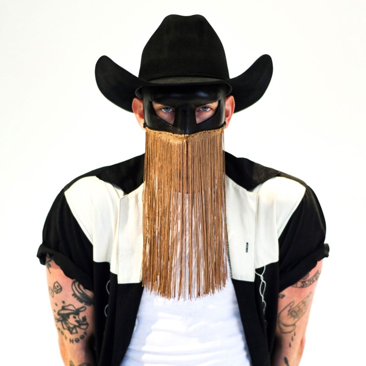 Orville Peck wearing black cowboy hat, eye mask with gold fringe, and black and white Western shirt